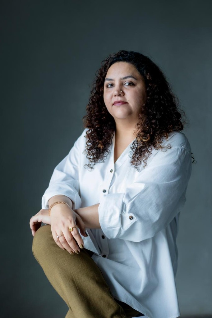 Professional portrait of Vrenda Kapoor, Narrative Architect and Creative Director, wearing a white button-down shirt against a dark grey background